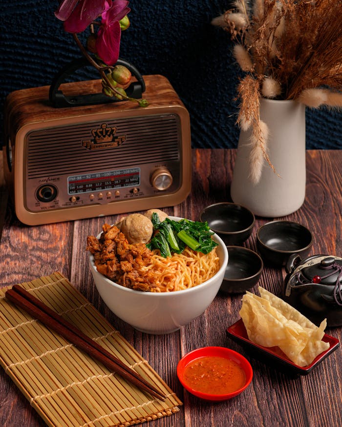 A vibrant noodle bowl with chicken and greens beside a vintage radio and classic decor.